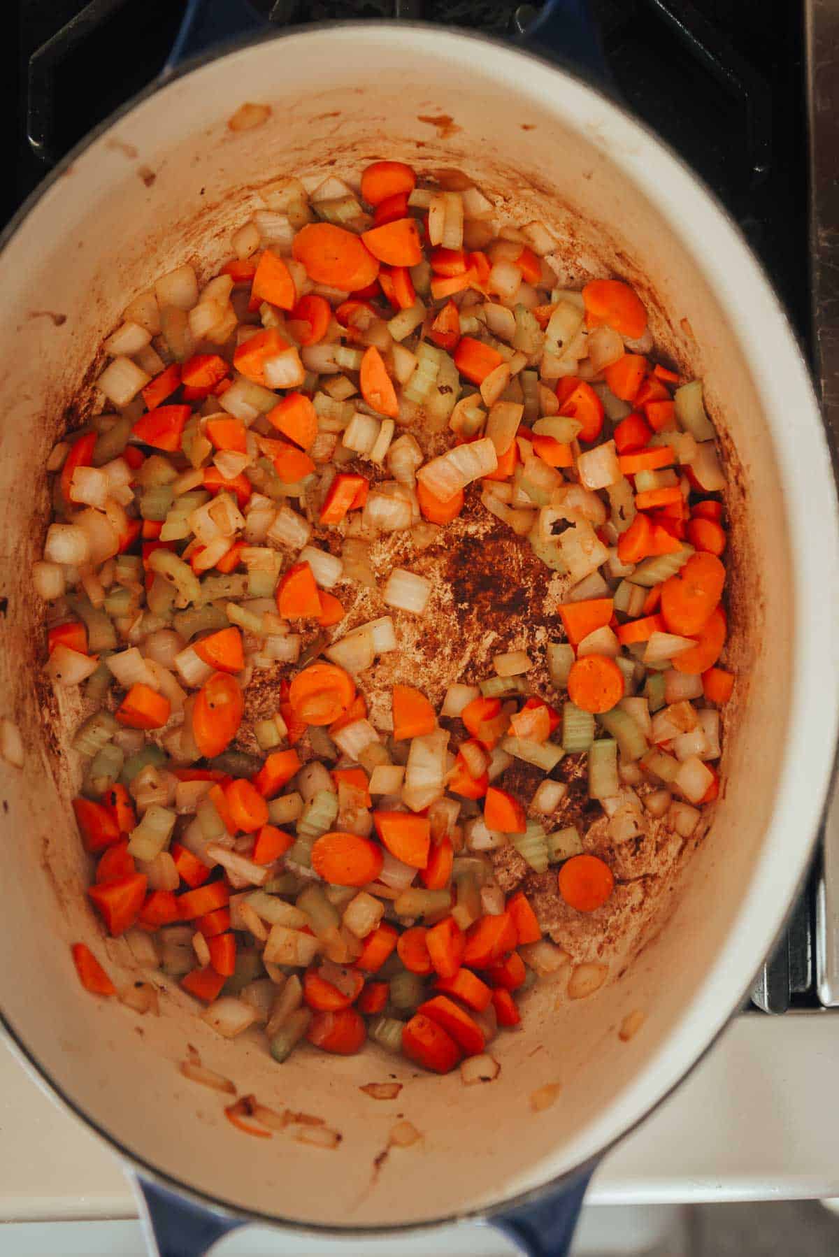 A pot on a stovetop containing sautéed diced carrots, onions, and celery.