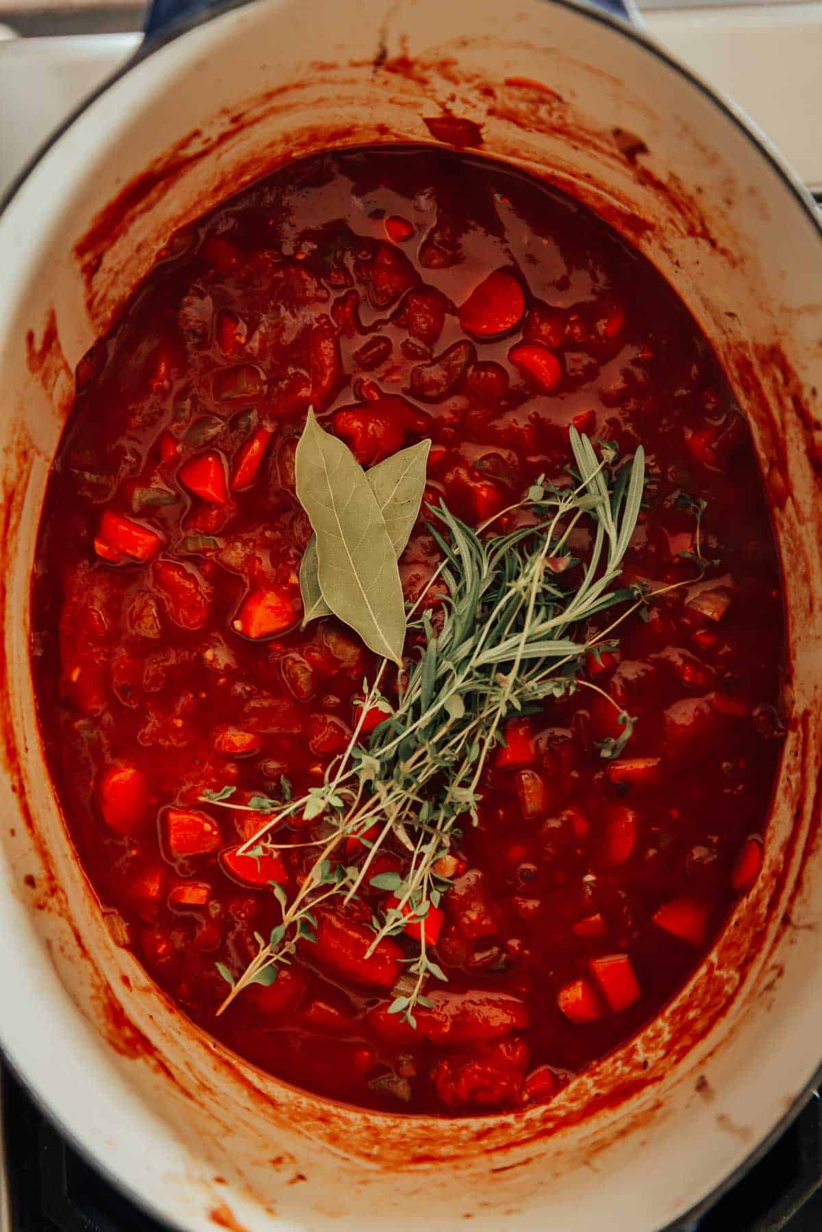 A pot of red stew with visible herbs including bay leaves and a sprig of thyme on top. The stew contains chunks of vegetables.