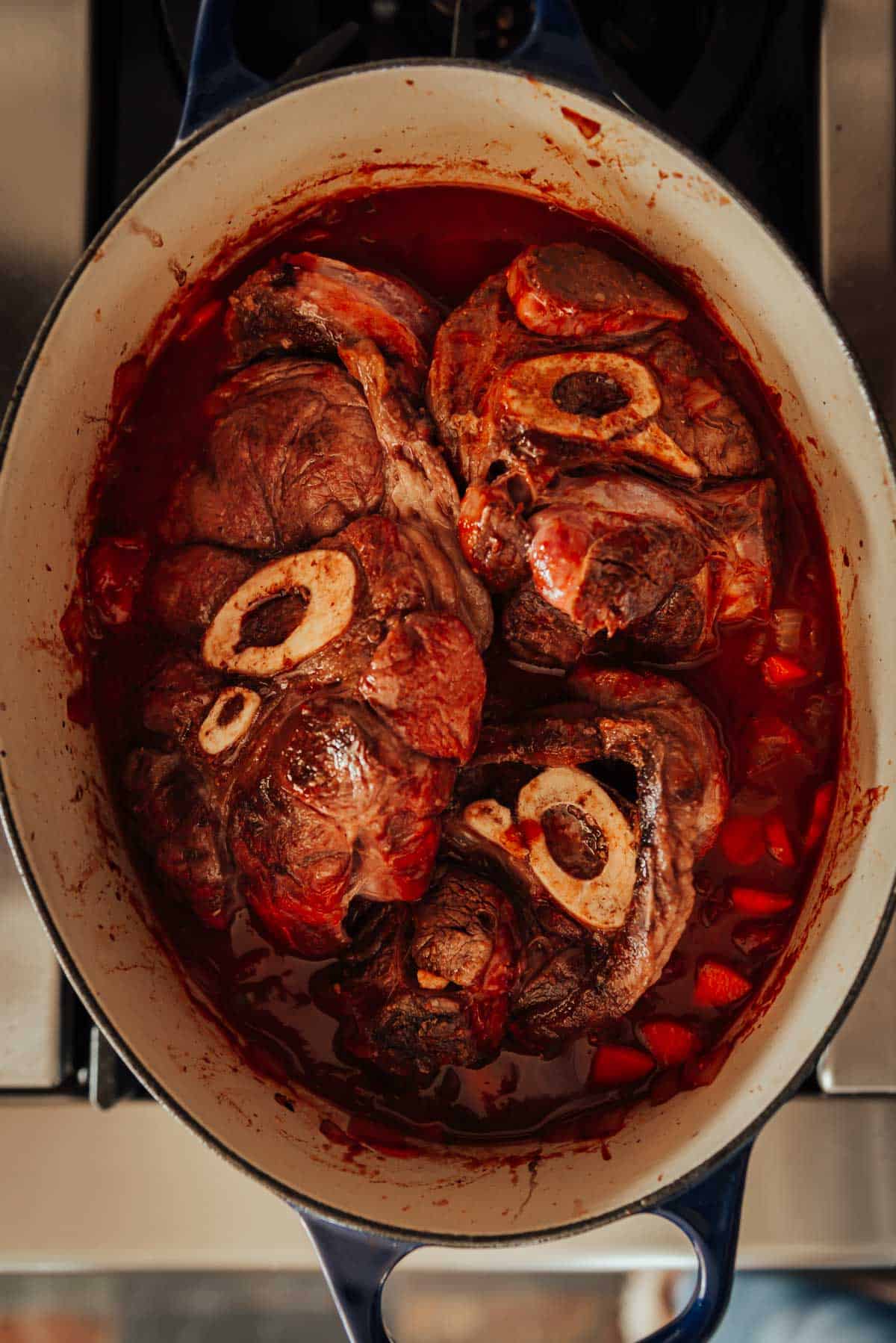 A pot filled with cooking meat and vegetables in a red sauce on a stove.