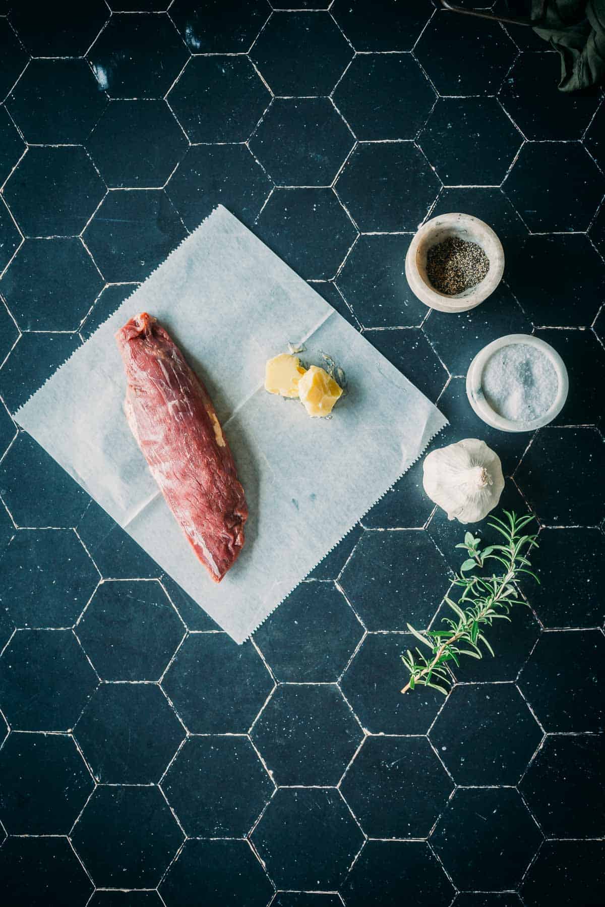 Overhead view of a raw beef cut on parchment paper, next to butter, salt, pepper, garlic, and a sprig of rosemary on a black hexagonal tile background.