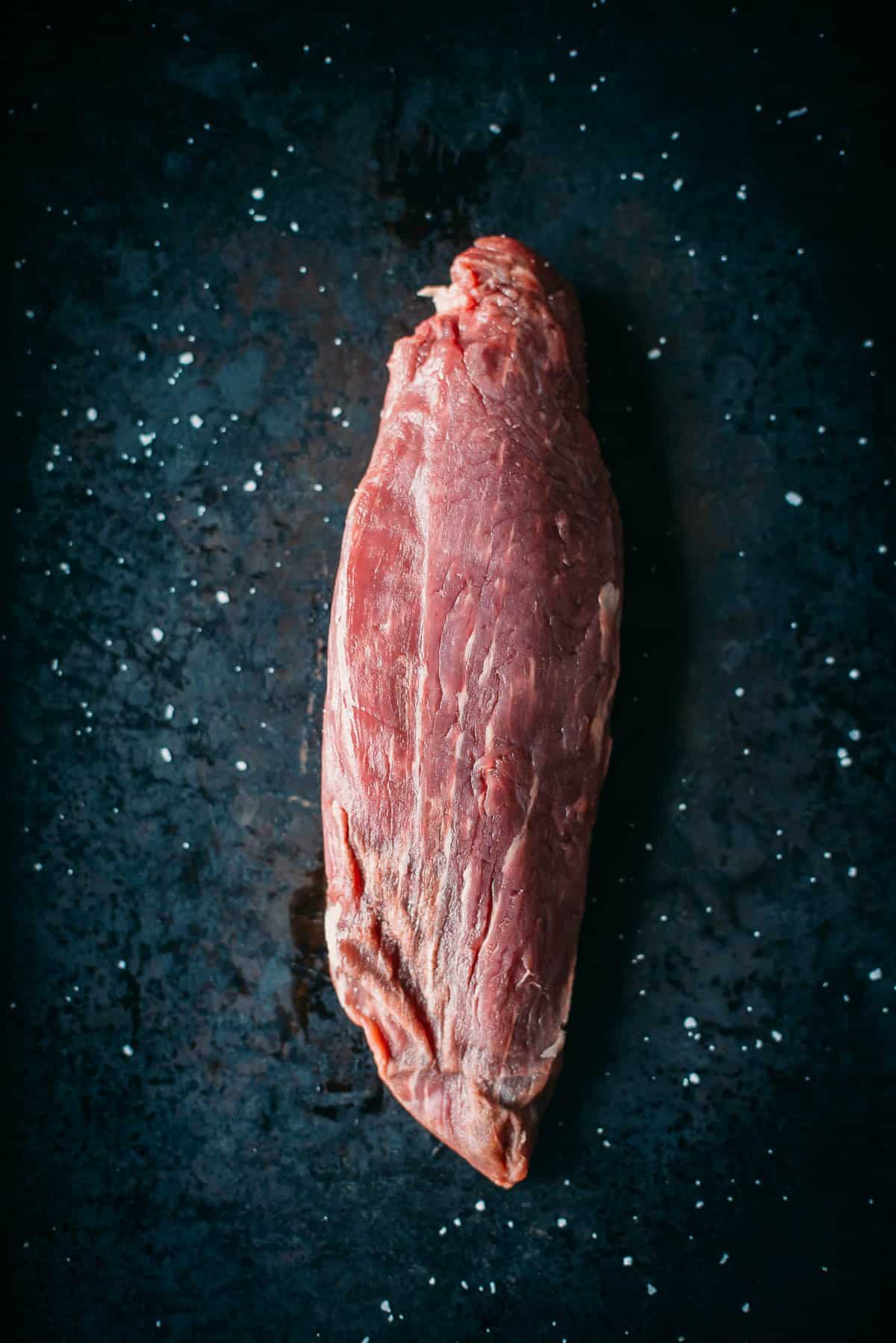 Raw teres major steak on a dark, speckled surface.