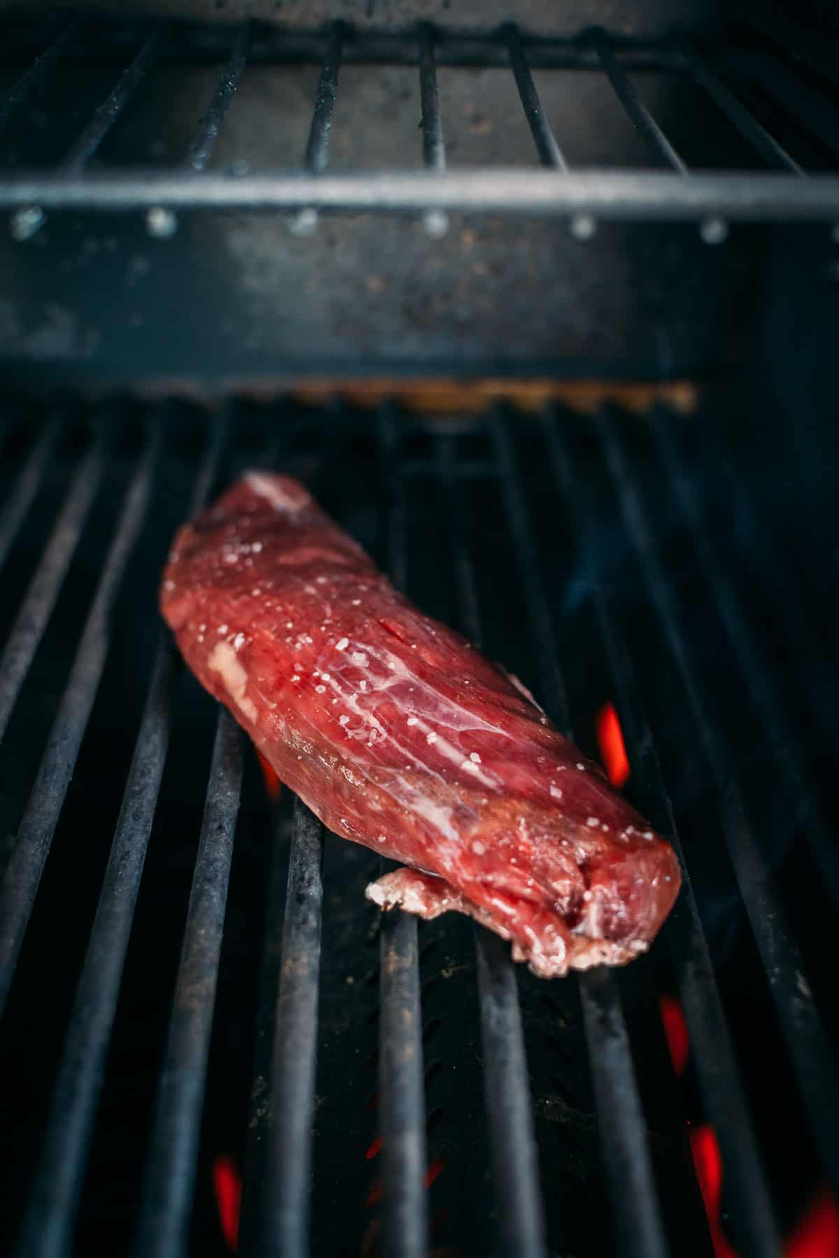 A piece of seasoned raw meat is placed on the grill grates, with visible flames underneath.