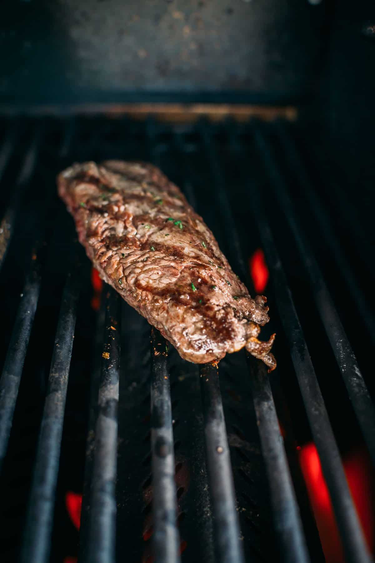 A piece of steak is being grilled on a barbecue grill with visible red hot coals beneath the grates.