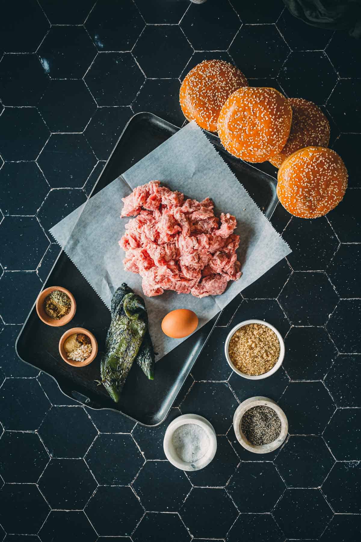 Overhead view of hamburger ingredients on a black tile surface, including ground meat, sesame seed buns, spices, an egg, and peppers on a tray.