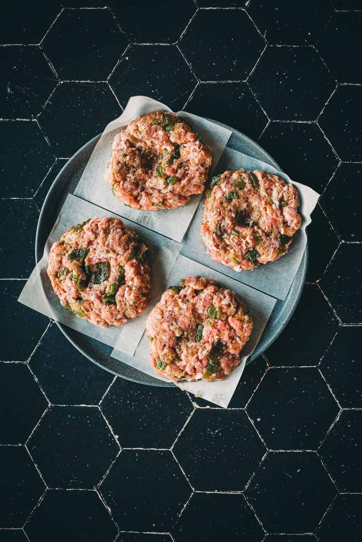 Four raw meat patties with chopped herbs on parchment paper, placed on a round plate on a black hexagonal tiled surface.