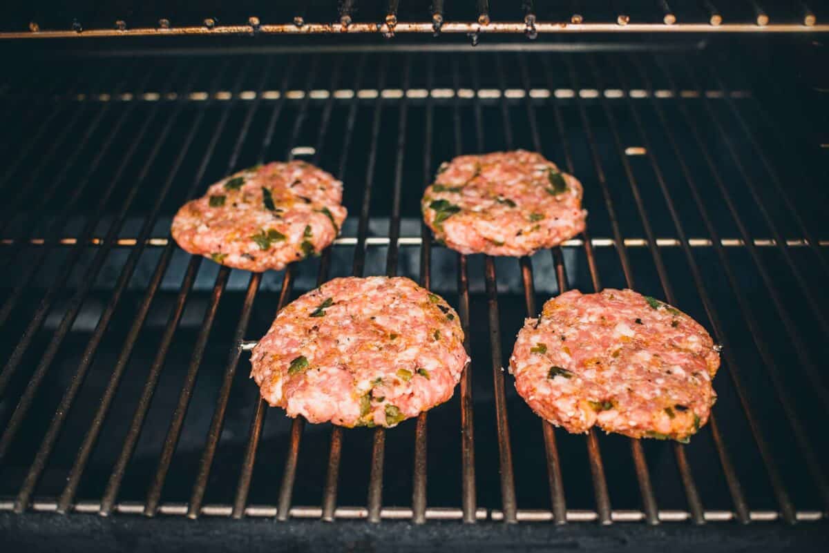 Four raw burger patties with visible green herbs are cooking on a grill.