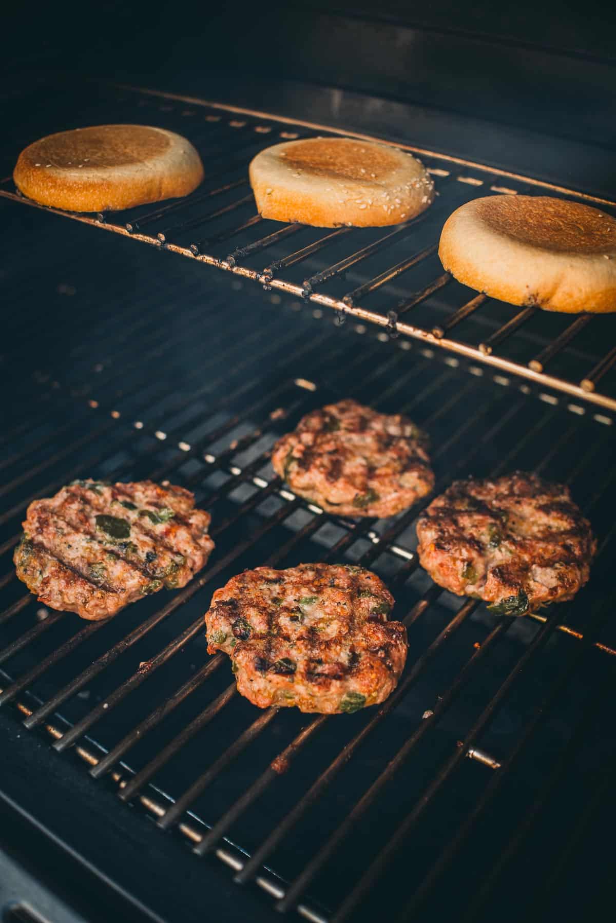 Four burger patties are being grilled on the bottom rack of a barbecue grill, while three burger buns toast on the top rack.