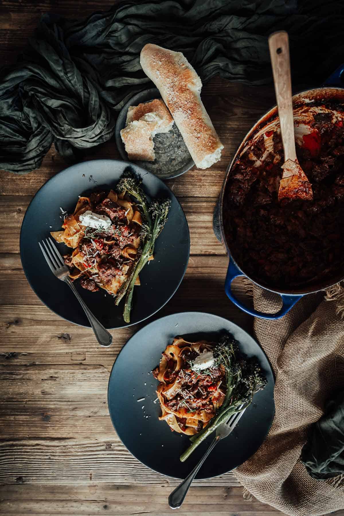 Two plates of pasta topped with osso buco and greens are set on a wooden table beside a loaf of bread. 