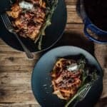 Two dark plates of pasta topped with osso buco and garnished with cheese and herbs, accompanied by broccolini and a fork next to each plate on a wooden table.