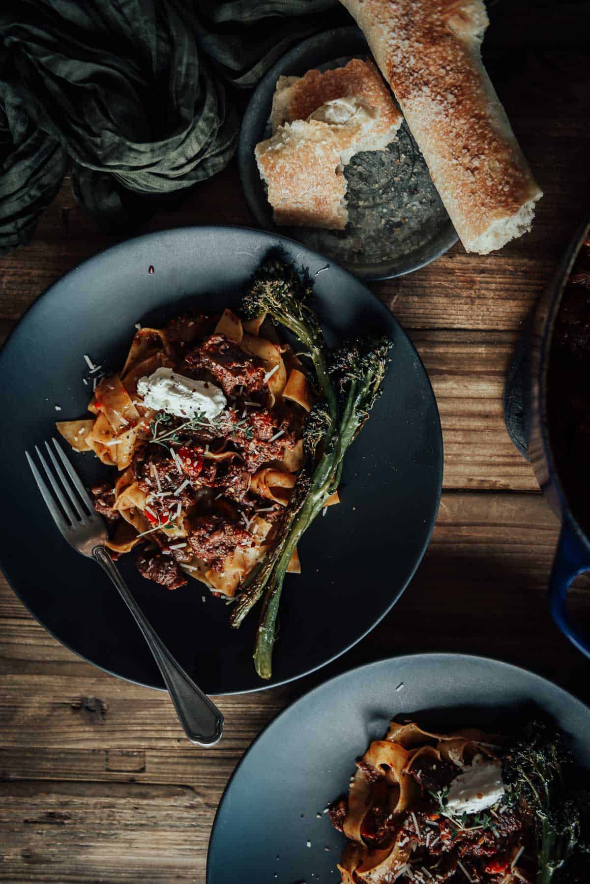 A wooden table with two plates of pasta topped with osso buco and a dollop of ricotta. 