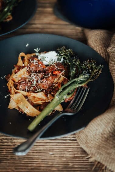 A plate of pappardelle pasta with osso buco, topped with a dollop of ricotta and grated cheese, served with a side of broccolini, and a fork on the side.