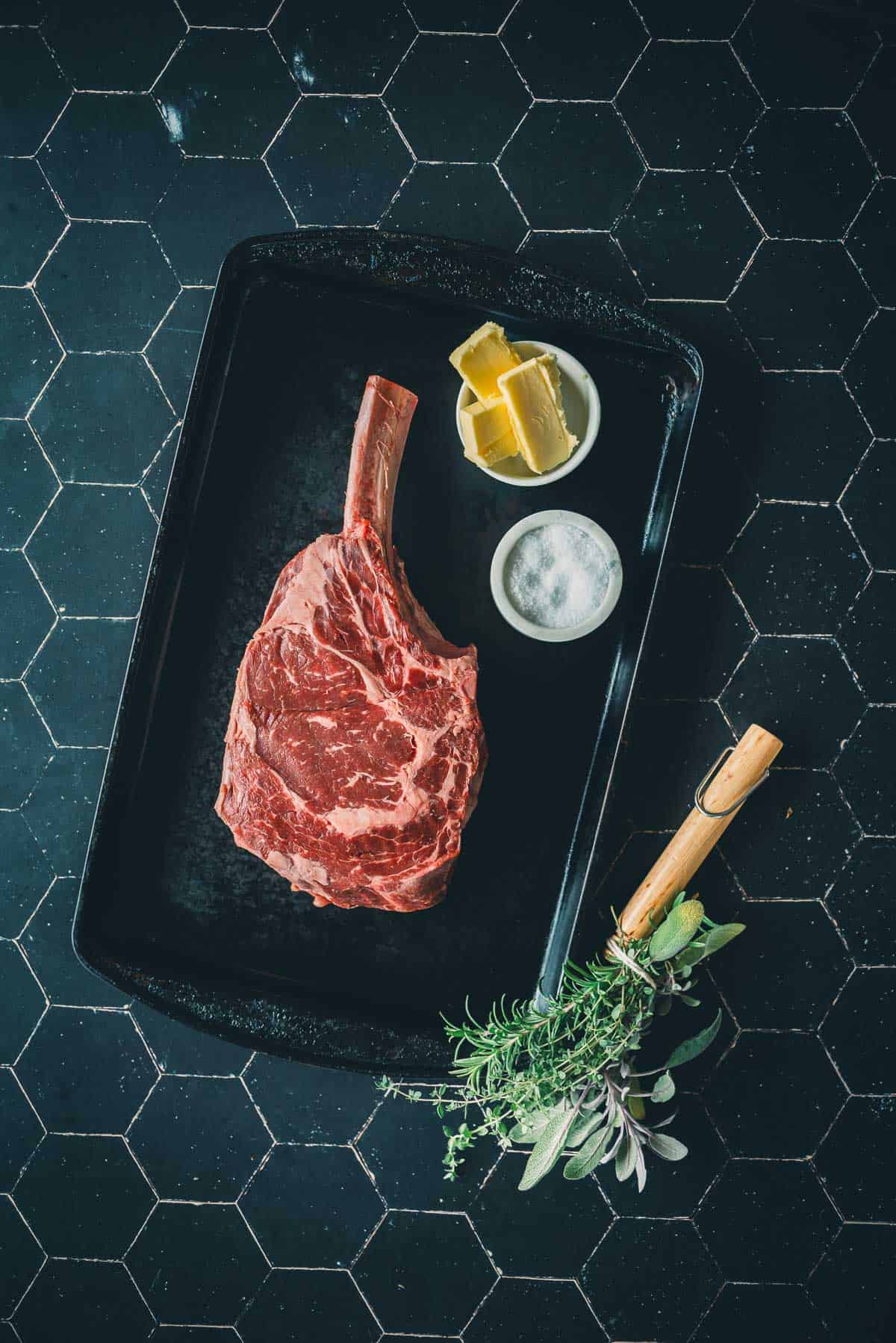 A raw cowboy ribeye steak on a tray with bowls of butter and salt beside it, and herbs bundled with a wooden handle placed nearby.