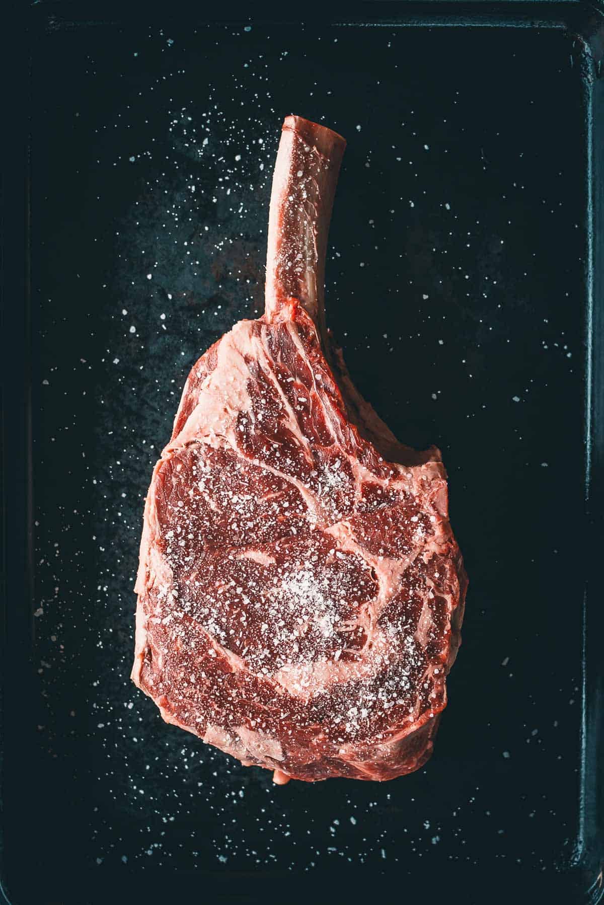 A raw, bone-in steak sprinkled with coarse salt on a dark surface.