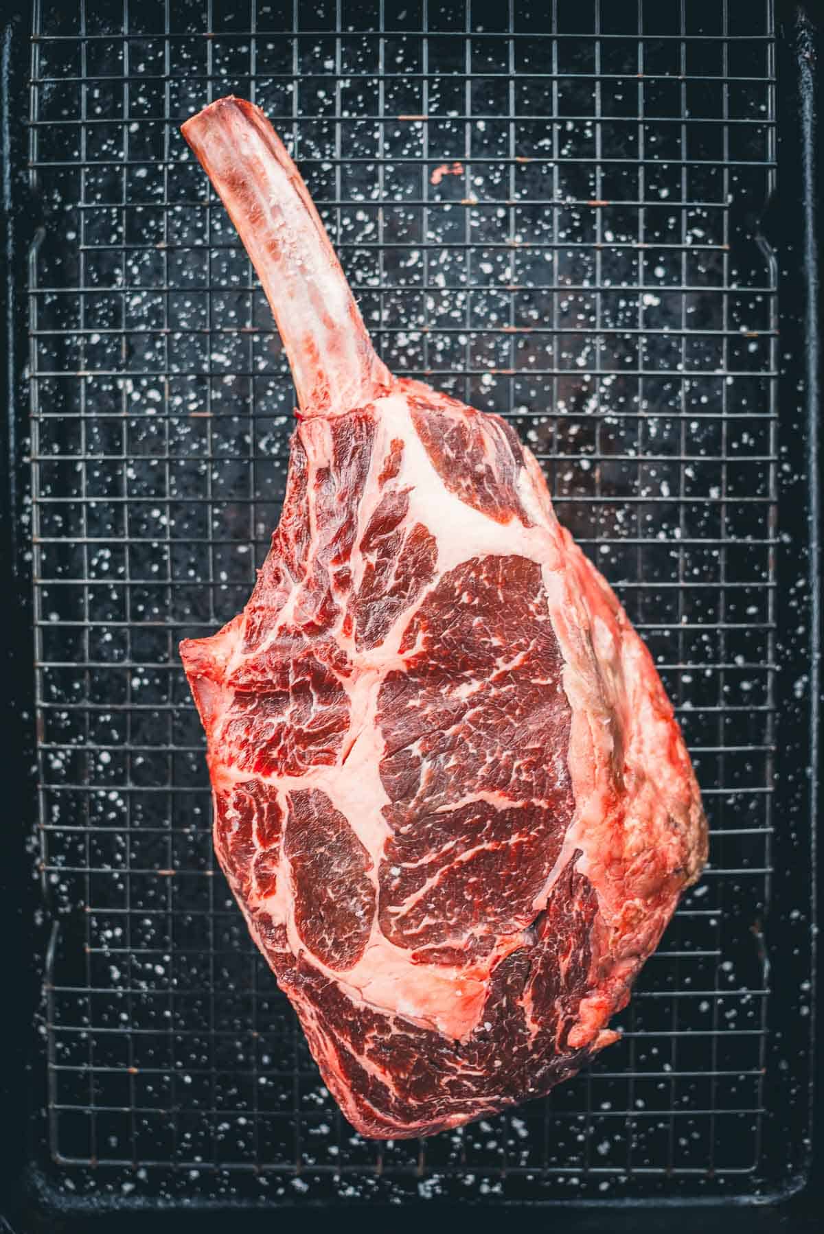 A raw tomahawk steak on a wire rack over a dark baking tray.