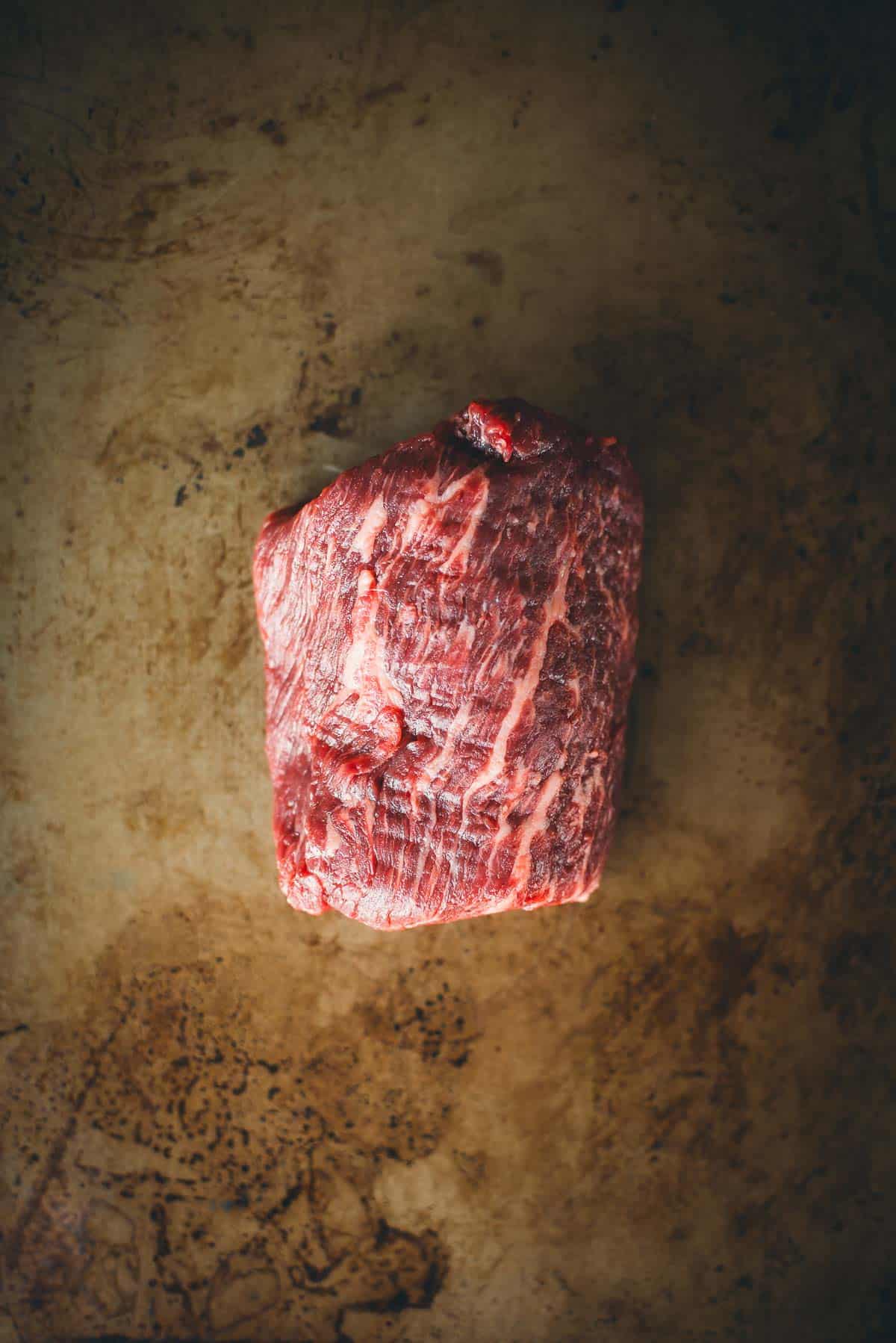 A raw piece of marbled Denver steak rests on a flat, textured brown surface.