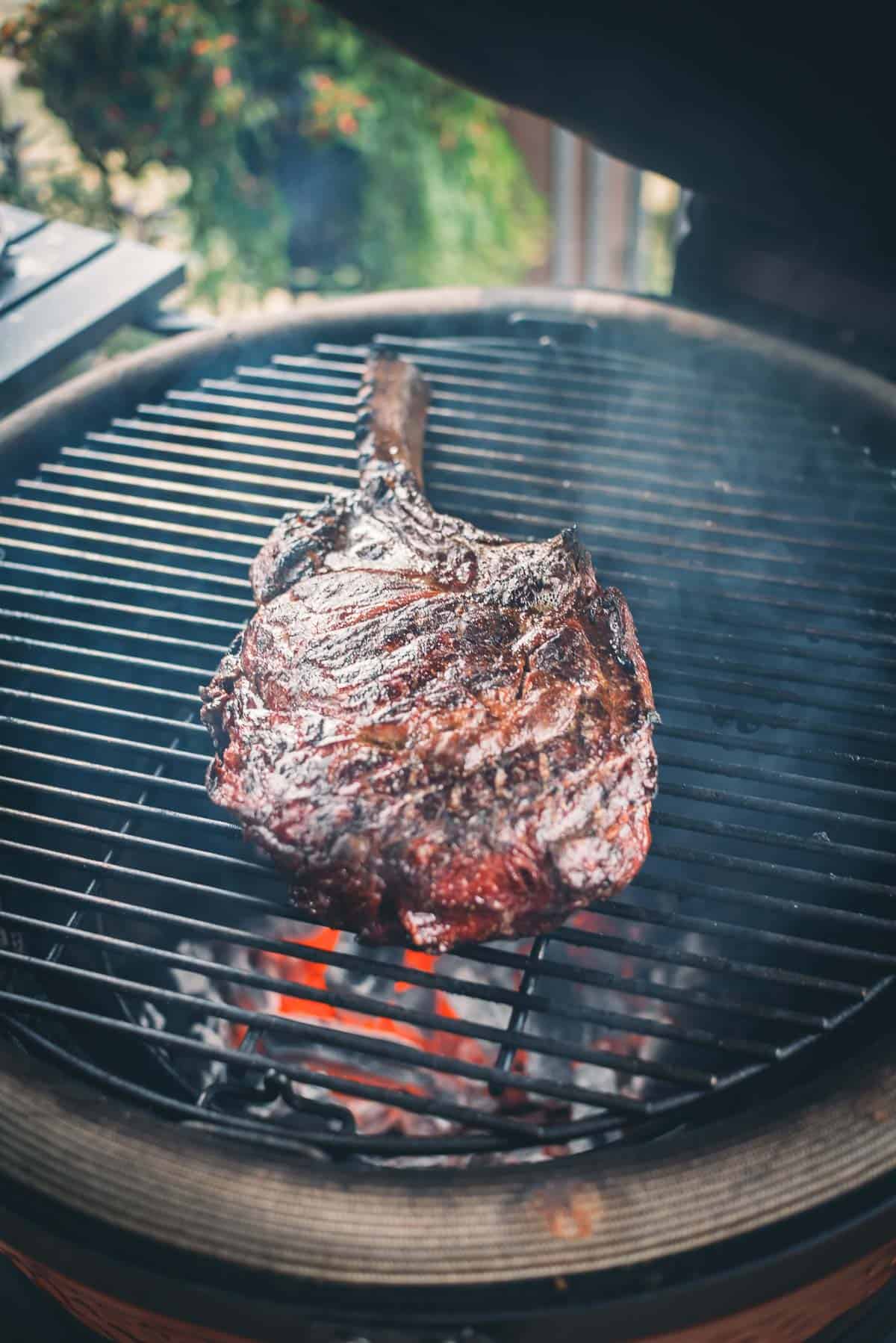 A large steak with a bone is cooking on a charcoal grill with visible orange coals underneath.