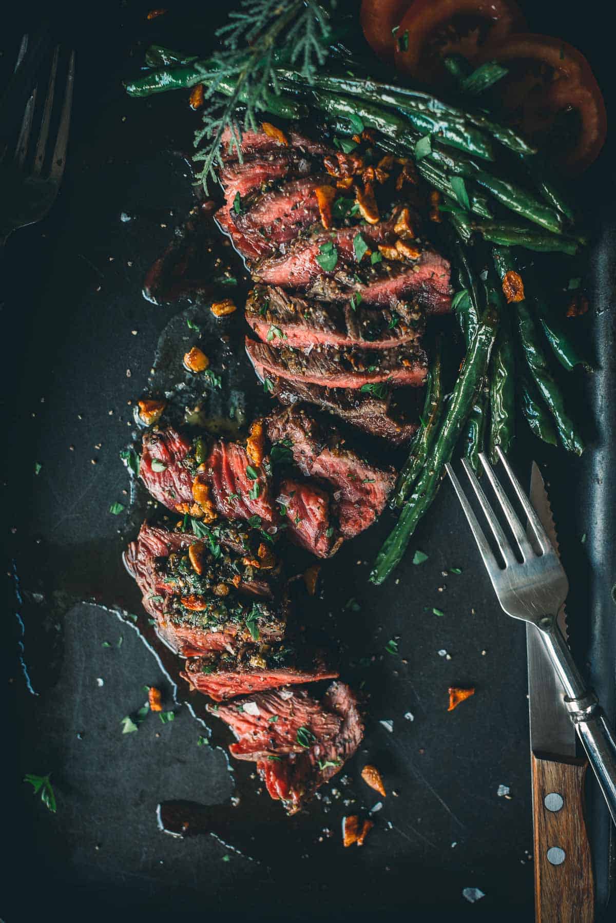 Sliced medium-rare steak garnished with herbs and garlic, accompanied by green beans and tomato slices, served on a dark plate with a fork and knife beside it.