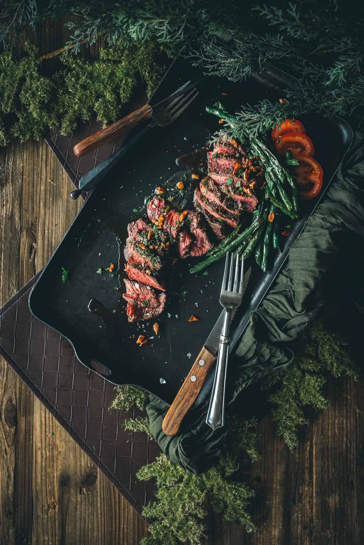 A serving of sliced medium-rare steak with green beans and tomato slices, garnished with herbs, on a dark tray with two wooden-handled steak knives and forks.