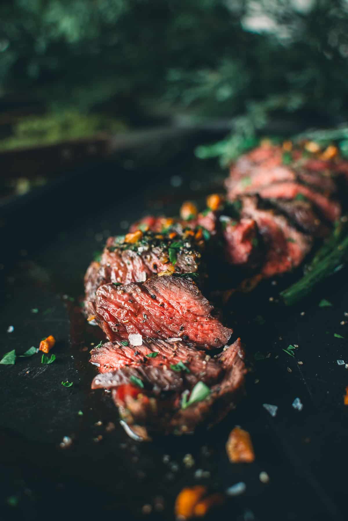 Close-up of sliced, medium-rare Denver steak on a dark surface, garnished with herbs and small pieces of vegetables.