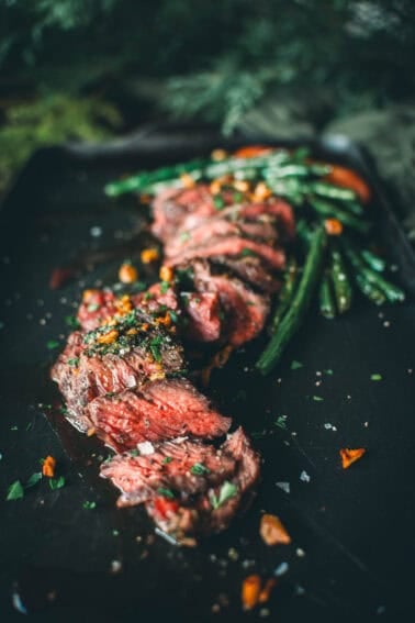 A sliced medium-rare steak garnished with herbs and coarse salt is served on a black plate alongside green beans and a few orange vegetables.