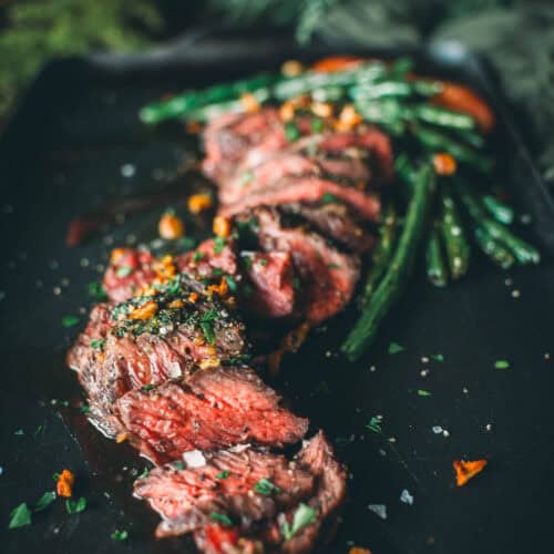 A sliced medium-rare steak garnished with herbs and coarse salt is served on a black plate alongside green beans and a few orange vegetables.