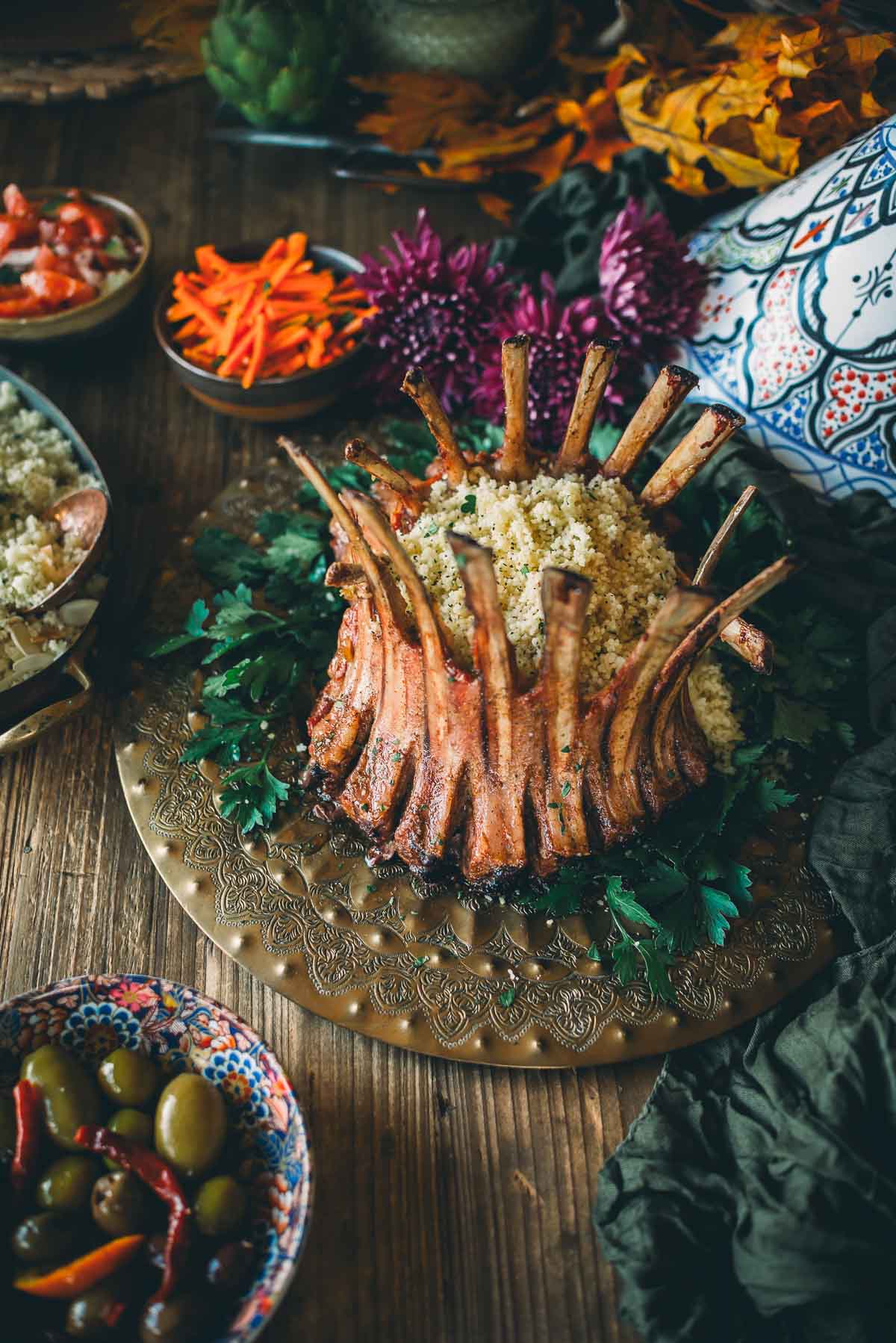 A roasted crown of lamb arranged in a crown shape with couscous in the center, served on a decorative platter with assorted sides, including olives and sliced vegetables, on a wooden table.