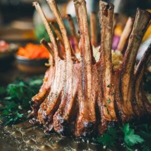 A close-up of a cooked crown roast of lamb garnished with parsley, displayed on an ornate platter with a blurred background of side dishes.