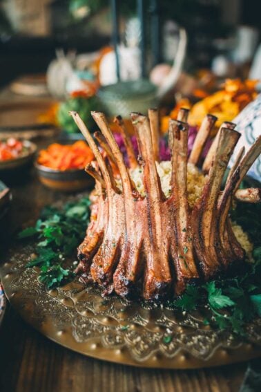 A perfectly cooked crown rack of lamb is served on a platter garnished with fresh greens, with sides of vegetables on a wooden table.