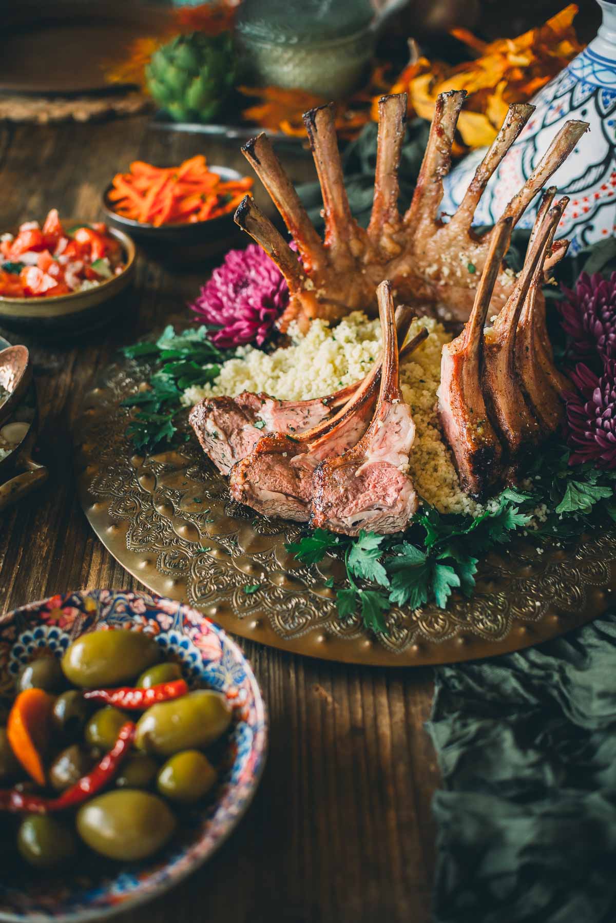 A plated meal featuring a lamb crown roast on a bed of couscous, accompanied by mixed olives, sliced carrots, and a floral garnish, is displayed on a decorated table.