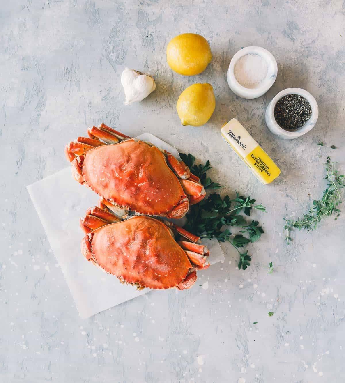 Two whole dungeness crabs on parchment paper with parsley, two lemons, garlic cloves, butter, salt, and pepper on a light gray surface.