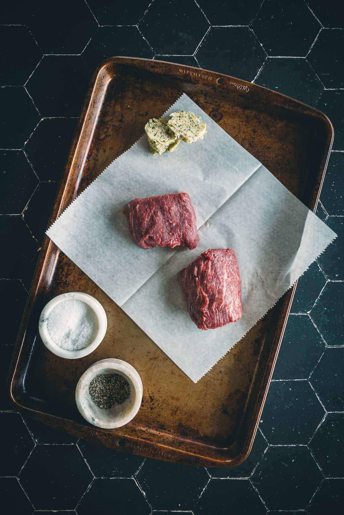 A baking tray with two raw denver steaks on parchment paper, two servings of herb butter, and small bowls of salt and pepper.