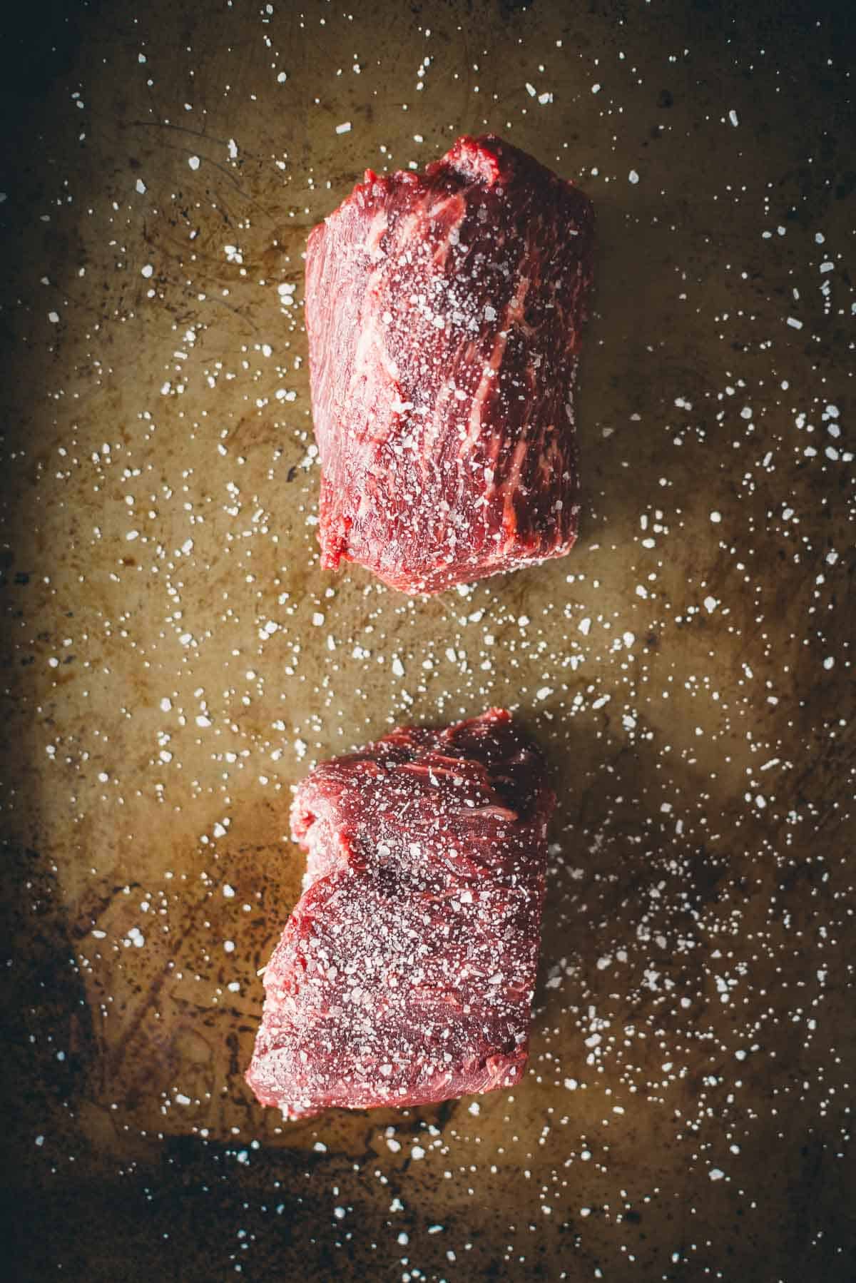 Two raw Denver steaks seasoned with salt on a brown surface.