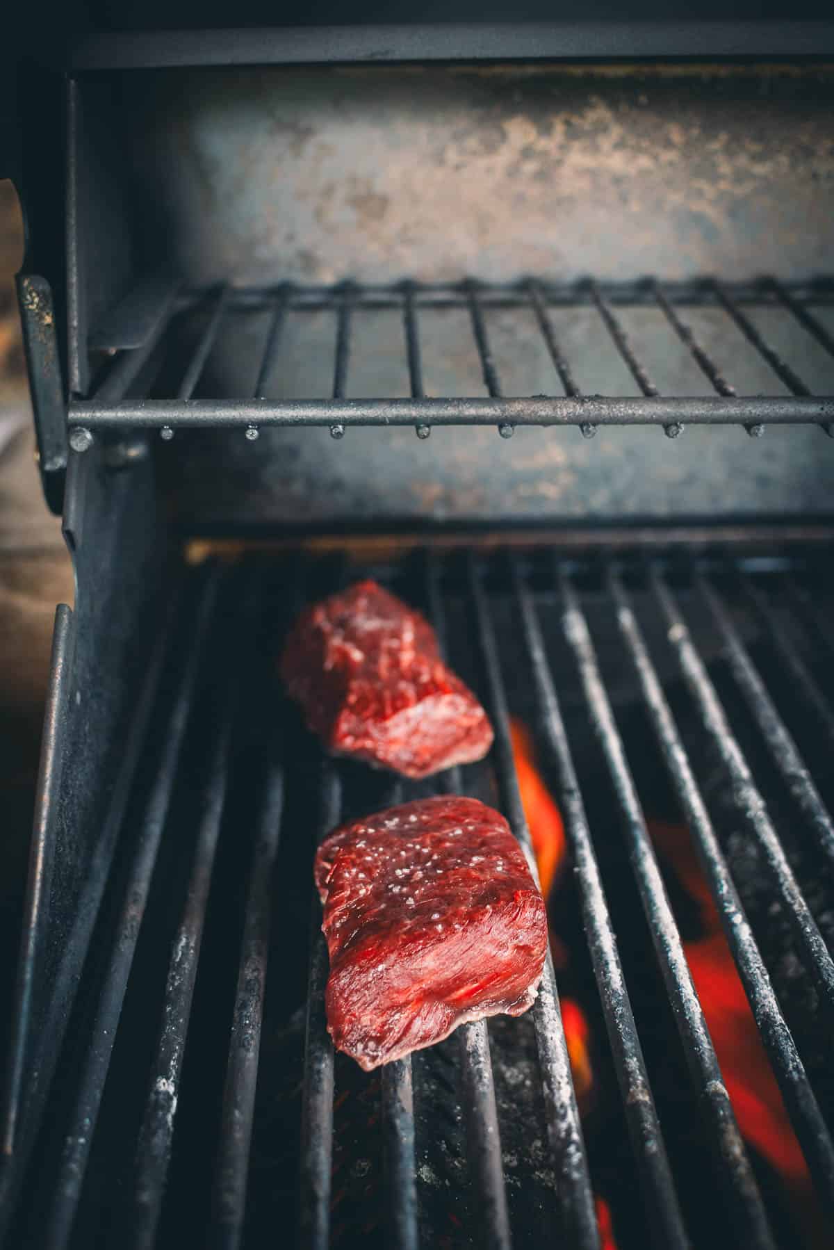 Two raw denver steaks placed on a grilling rack with visible flames below on a barbecue grill.