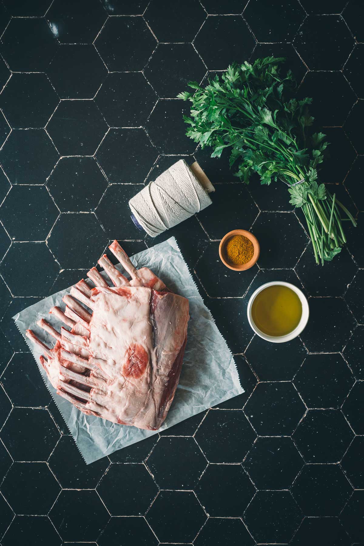 Raw rack of lamb on parchment paper, a bunch of fresh parsley, a small bowl of spices, a cup of olive oil, and kitchen twine on a black hexagon-tiled countertop.