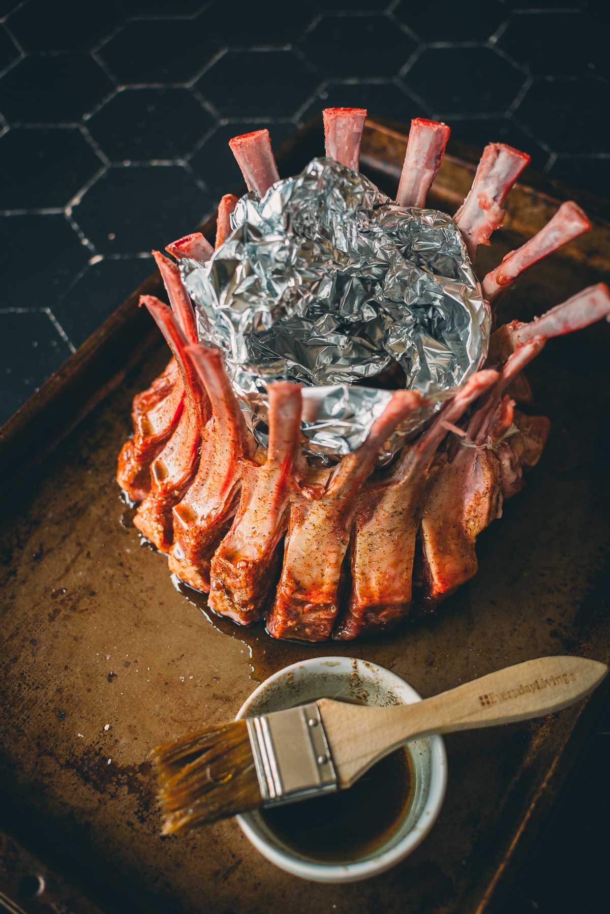 A raw crown roast of lamb with rib bones wrapped in aluminum foil sits on a baking sheet, next to a small bowl of marinade and a basting brush.