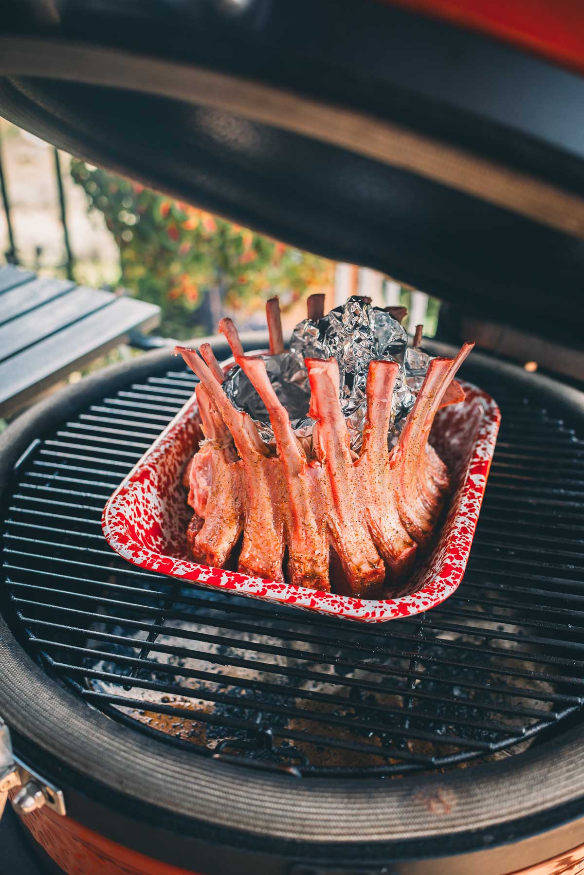 A rack of seasoned meat ribs arranged in a circular pattern on a grill, cooking in a covered red roasting pan.