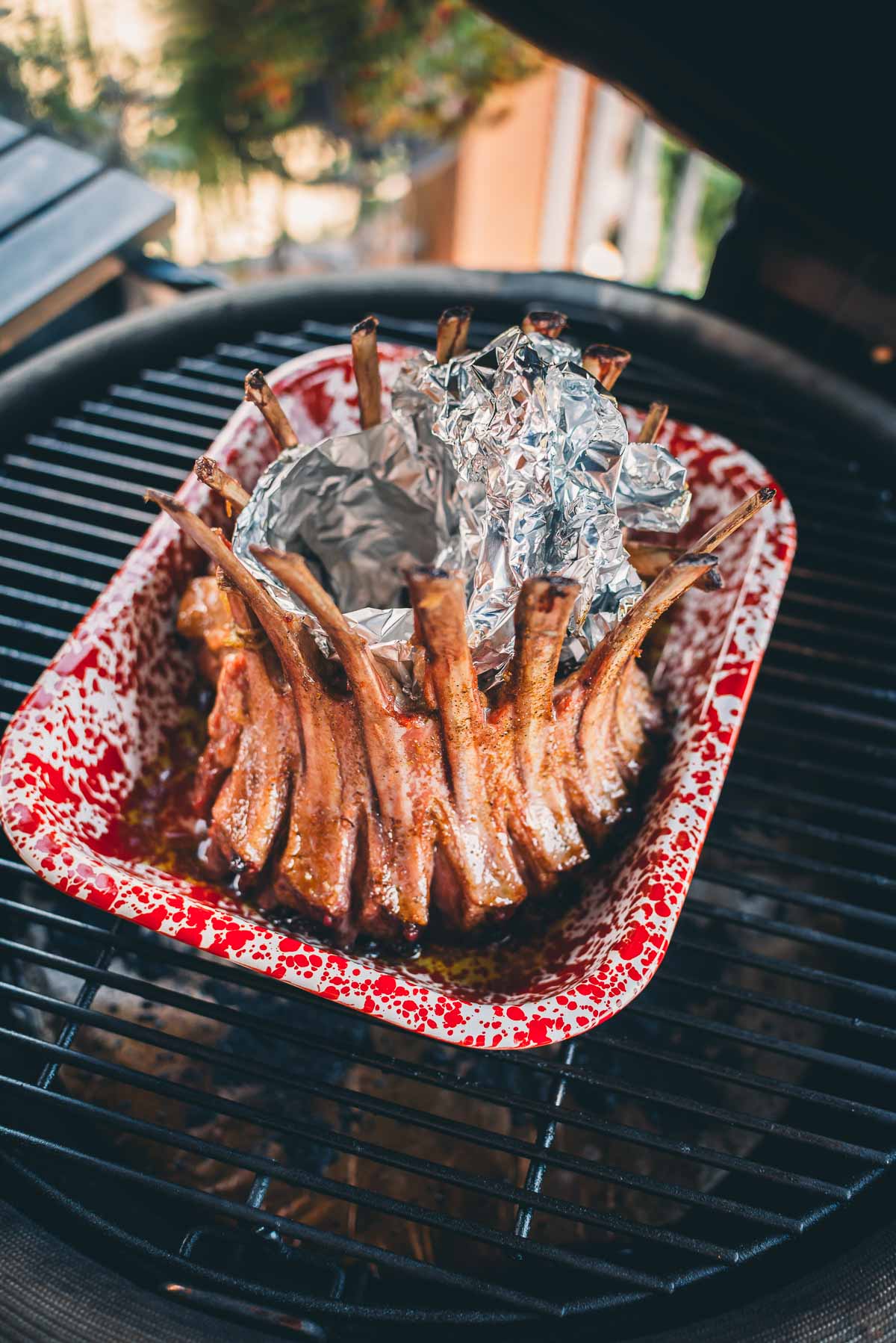 A crown roast of lamb wrapped in foil in a red and white speckled roasting pan on a grill.