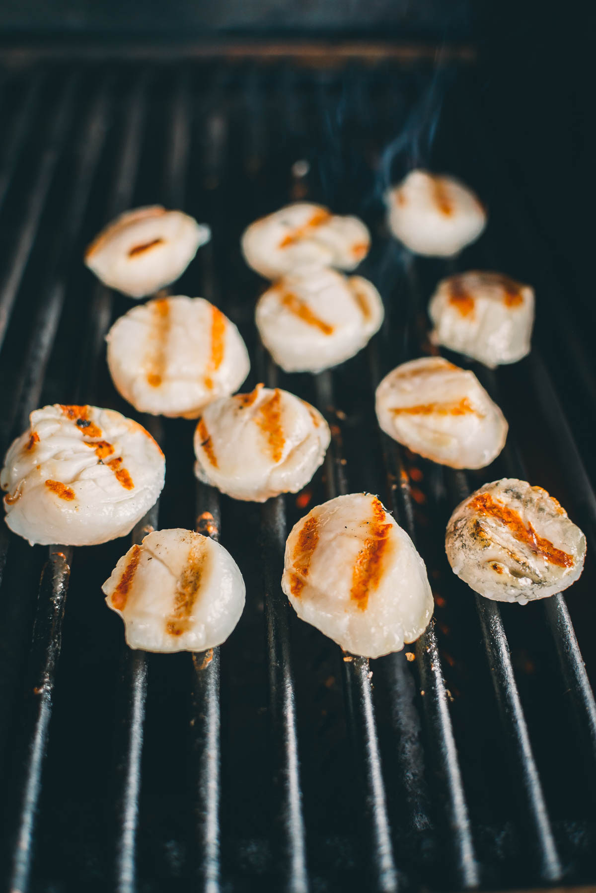 Scallops grilling on a barbecue, with visible sear marks and small wisps of smoke rising from some of them.