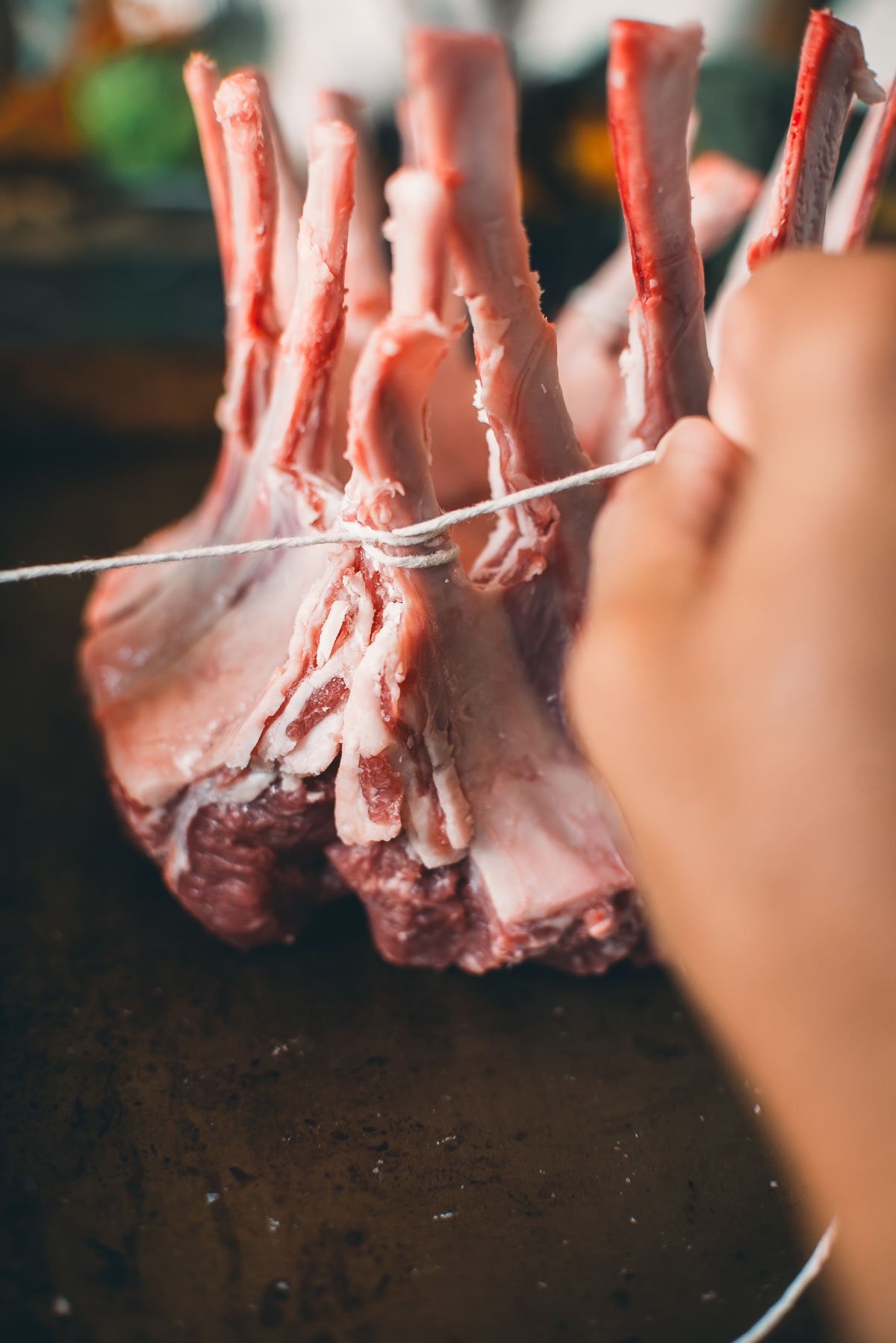 A hand is tying a string around a raw rack of lamb standing on a dark surface.