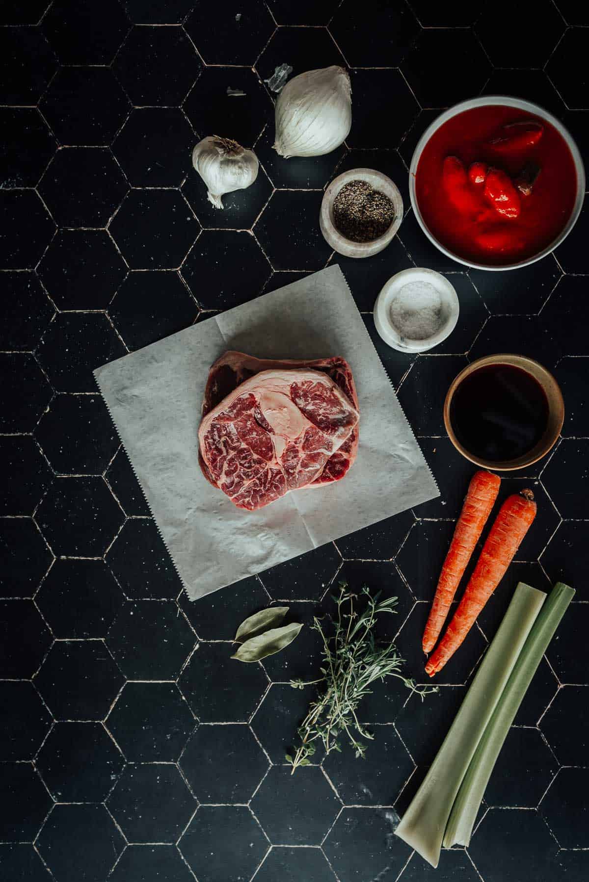 Cooking ingredients arranged on a black hexagonal tile surface, including a raw beef shank, carrots, celery, garlic, onion, bay leaves, herbs, tomatoes in a bowl, salt, pepper, and a bowl of sauce.