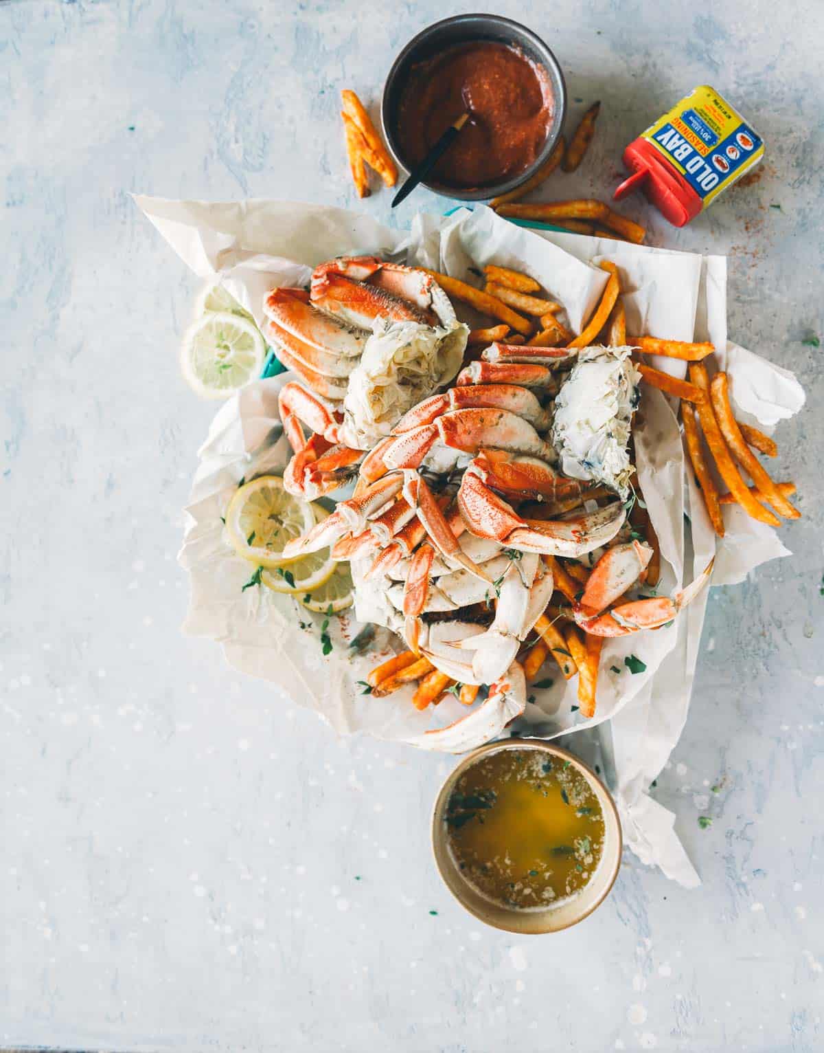 A dish consisting of crab legs, French fries, lemon slices, and two dipping sauces is arranged on paper with a blue-gray background.