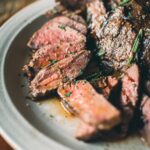 A plate of sliced medium-rare elk steaks garnished with fresh rosemary is served on a table.