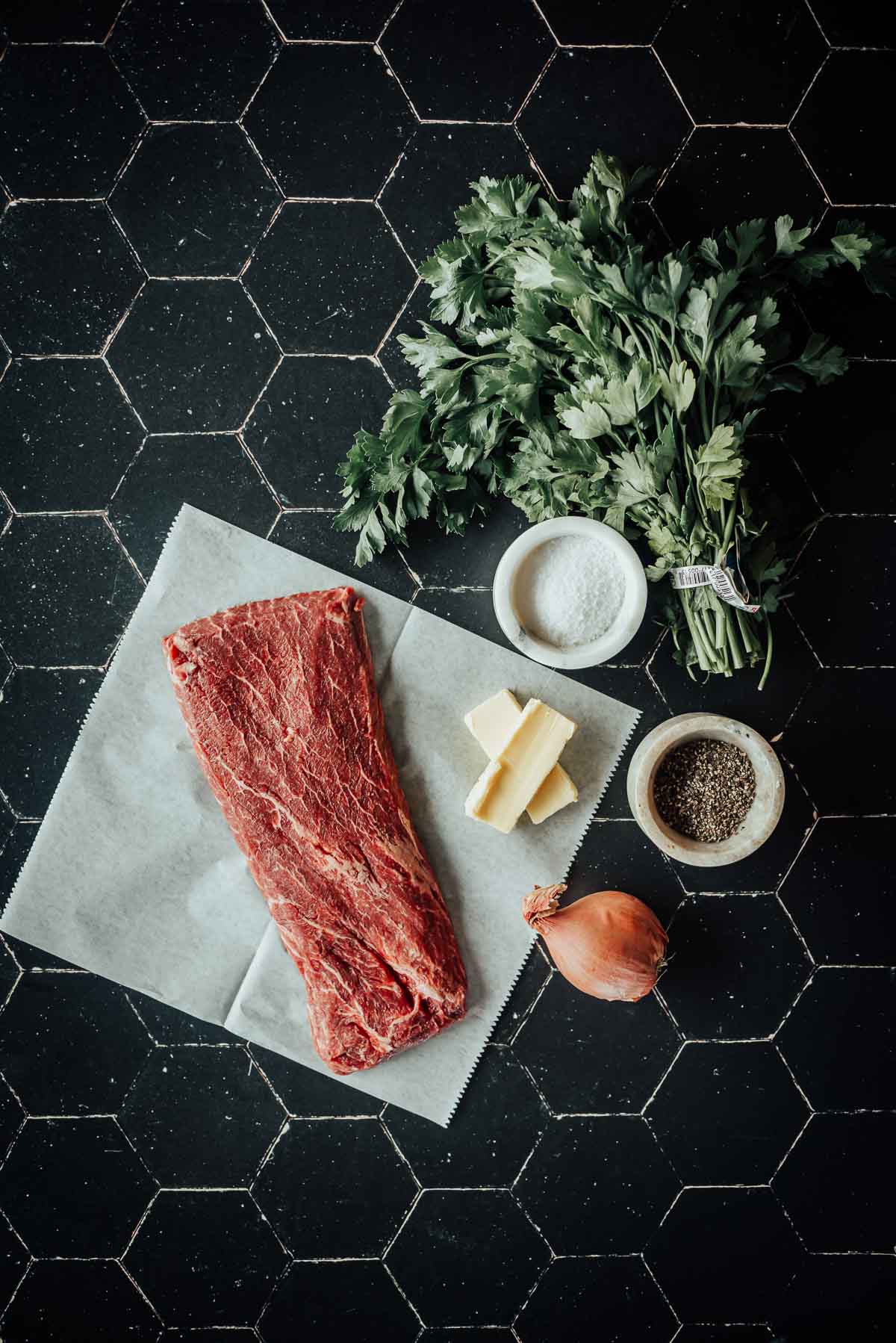 A raw flat iron steak, parsley, butter, salt, pepper, and a shallot are arranged on a black tiled surface.