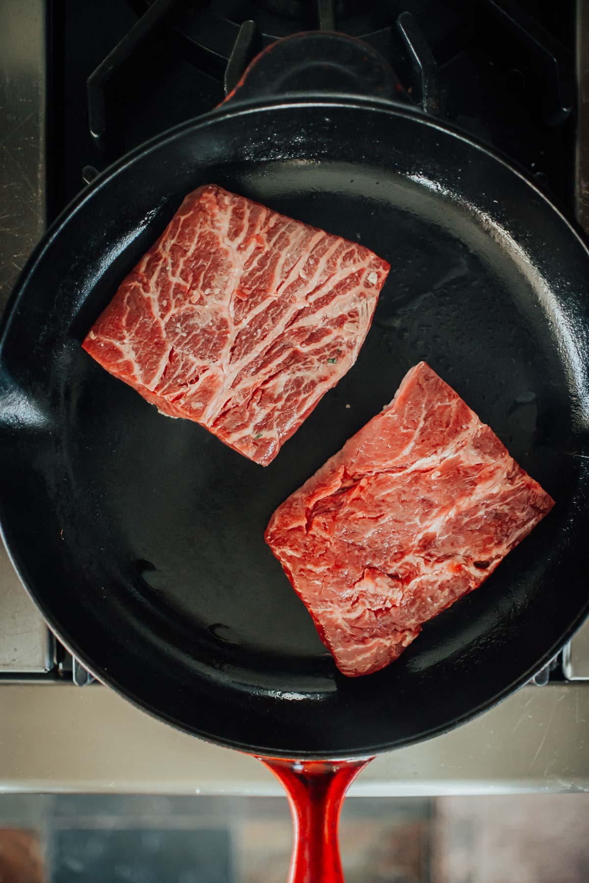 Two raw marbled flat iron steaks are placed in a black skillet on a stovetop.