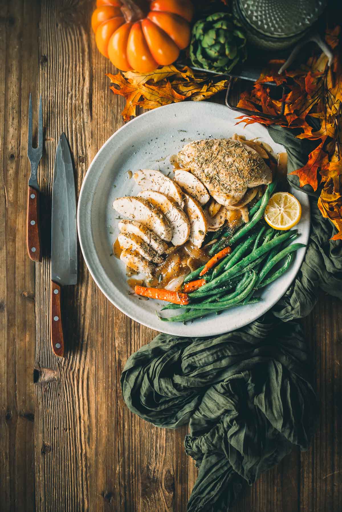 Plate of sliced roast turkey tenderloins with green beans and carrots, garnished with herbs, lemon, and accompanied by a carving knife and fork set on a wooden table with autumn decorations.