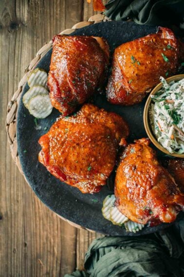 A plate of smoked turkey thighs, garnished with herbs, pickles on the side, and a small bowl of coleslaw.