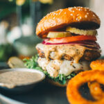 A turkey burger with lettuce, tomatoes, pickles, onions, melted cheese, and sauce on a sesame seed bun, served alongside a dipping sauce and onion rings.