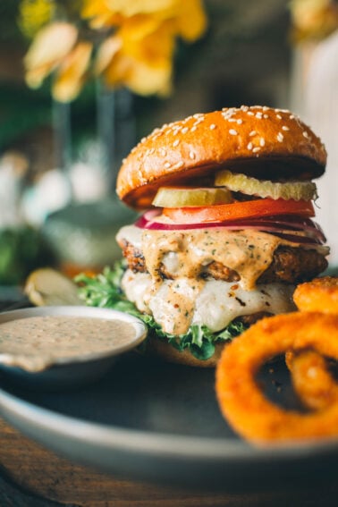 A turkey burger with lettuce, tomatoes, pickles, onions, melted cheese, and sauce on a sesame seed bun, served alongside a dipping sauce and onion rings.