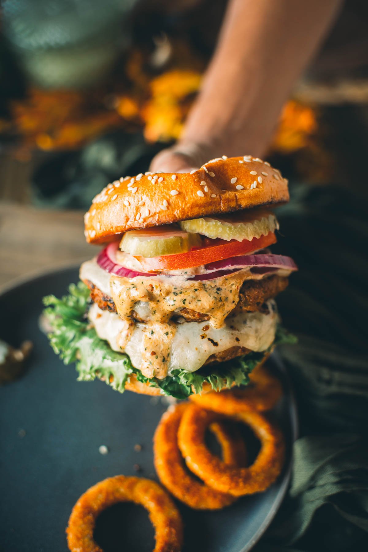 Close-up of a hand holding a juicy turkey burger with lettuce, tomato, onion, pickles, melted cheese, and dripping with sauce in a sesame seed bun. 