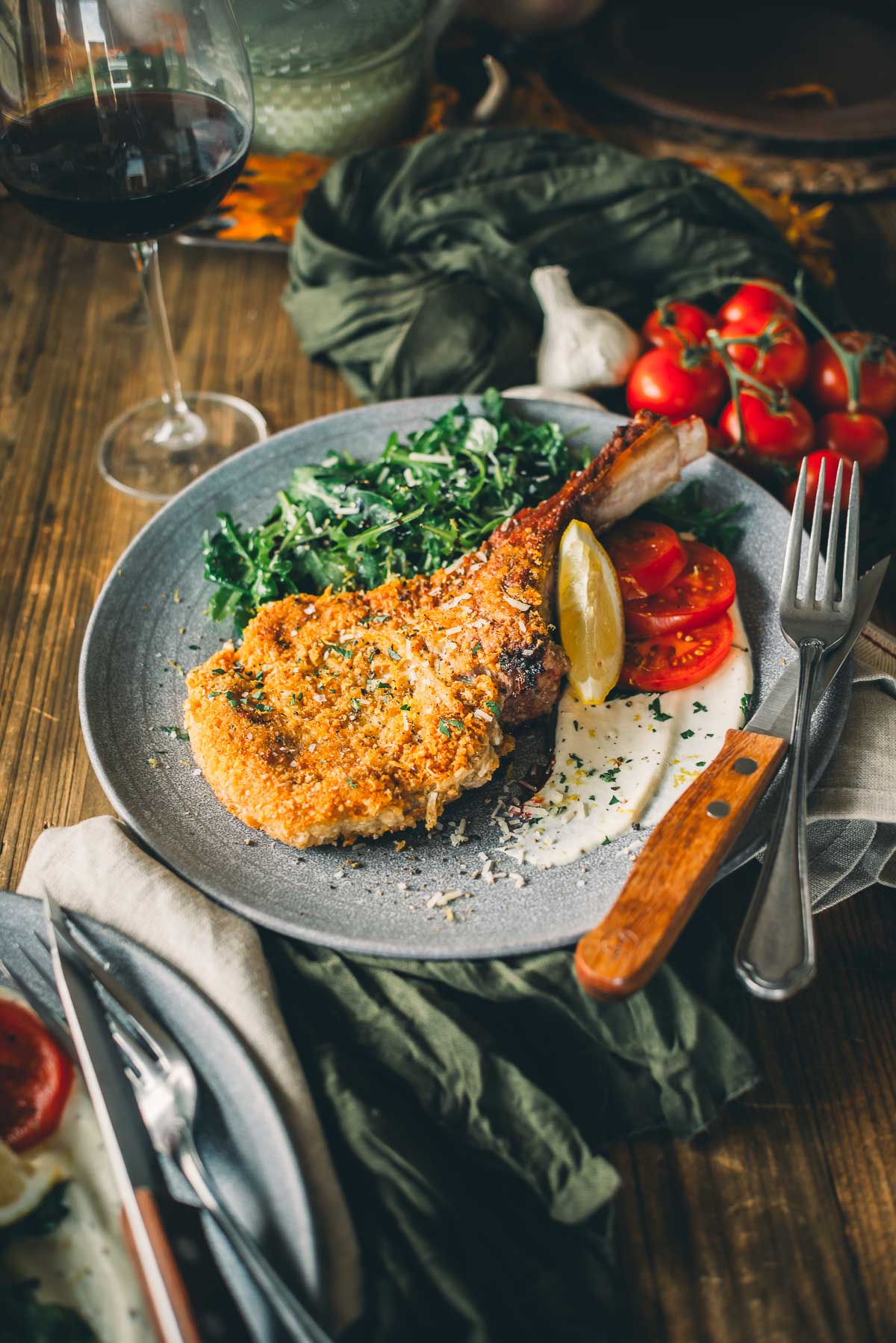 Breaded veal milanese with a lemon wedge, tomatoes, and greens on a gray plate, accompanied by a fork and knife, with a glass of red wine in the background.